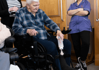 Resident in a wheelchair interacting with a visiting therapy animal while care staff offer support.