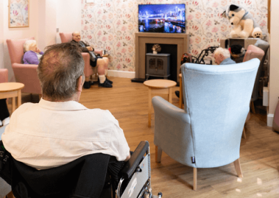 Residents seated in a cosy communal lounge area watching television, with comfortable armchairs, patterned wallpaper and wooden flooring.