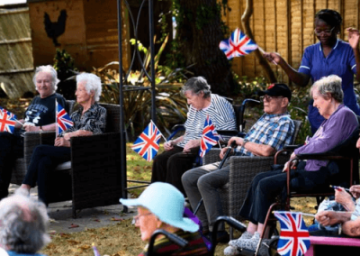 Residents seated outdoors holding Union Jack flags during a garden celebration with care staff present.