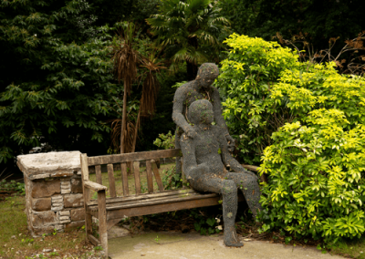 Decorative garden sculpture of two figures seated on a bench surrounded by lush plants and trees.