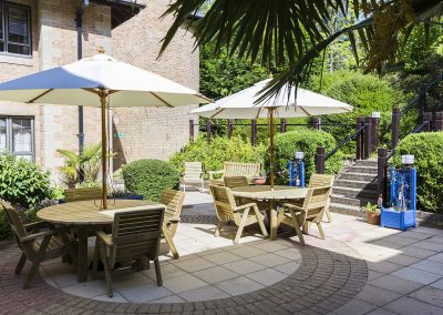 Patio terrace with wooden tables, chairs and parasols set within landscaped garden surroundings.